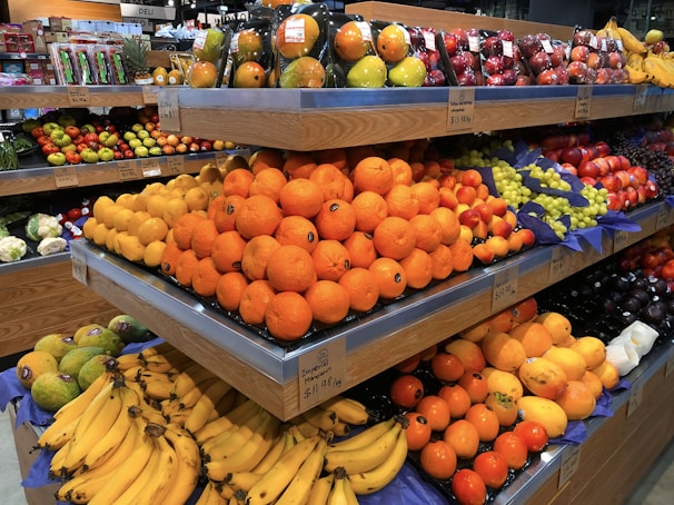 A supermarket produce section with stacked shelves displaying a variety of fresh fruits including oranges, bananas, grapes, and apples. The fruits are arranged neatly with prices labeled below each section.