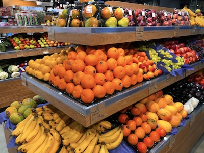 A supermarket produce section with stacked shelves displaying a variety of fresh fruits including oranges, bananas, grapes, and apples. The fruits are arranged neatly with prices labeled below each section.