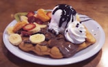 A delectable dessert featuring a bubble waffle topped with a scoop of vanilla ice cream drizzled with chocolate sauce. The waffle is garnished with an assortment of fruits including sliced strawberries, bananas, kiwi, and orange segments. A dollop of whipped cream adds to the indulgence.