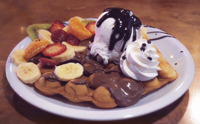 A delectable dessert featuring a bubble waffle topped with a scoop of vanilla ice cream drizzled with chocolate sauce. The waffle is garnished with an assortment of fruits including sliced strawberries, bananas, kiwi, and orange segments. A dollop of whipped cream adds to the indulgence.