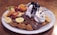 A delectable dessert featuring a bubble waffle topped with a scoop of vanilla ice cream drizzled with chocolate sauce. The waffle is garnished with an assortment of fruits including sliced strawberries, bananas, kiwi, and orange segments. A dollop of whipped cream adds to the indulgence.