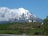 A picturesque snow-capped mountain rises prominently in the background, with a modern house situated on a lush green hillside in the foreground. Cattle are grazing on the expansive grassland that covers the hillside. Clouds partially obscure the peak of the mountain, adding to the serene and natural setting.