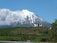 A picturesque snow-capped mountain rises prominently in the background, with a modern house situated on a lush green hillside in the foreground. Cattle are grazing on the expansive grassland that covers the hillside. Clouds partially obscure the peak of the mountain, adding to the serene and natural setting.
