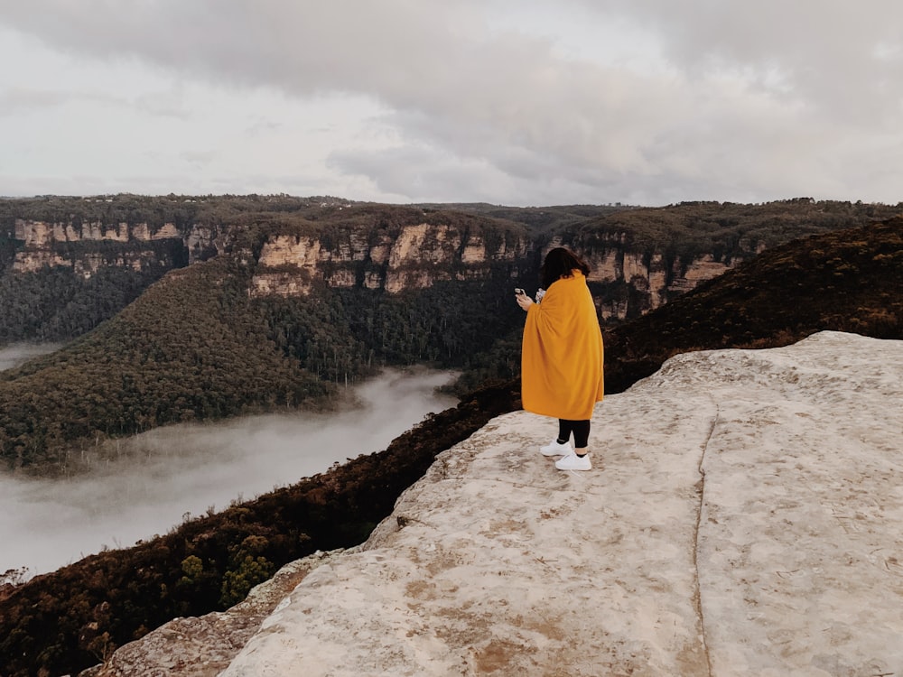 woman in yellow hoodie standing on rock formation near body of water during daytime