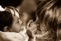A small child and an adult are face-to-face, engaging in a close, intimate interaction. The child looks intently at the adult, who is smiling warmly, creating a gentle and loving atmosphere. The image is in sepia tones, adding a nostalgic feel.