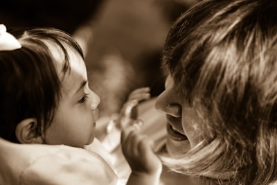 A small child and an adult are face-to-face, engaging in a close, intimate interaction. The child looks intently at the adult, who is smiling warmly, creating a gentle and loving atmosphere. The image is in sepia tones, adding a nostalgic feel.