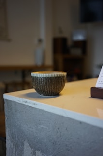 A textured ceramic cup with a ribbed design rests on a smooth, off-white countertop. The background appears blurred, giving an out-of-focus view of possible furniture or decor, contributing to a cozy and minimalist ambiance.
