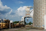 An industrial setting featuring a large, cylindrical metal silo with rust and a conveyor structure attached to it. A couple of pieces of heavy machinery are parked nearby, and a sign warns about keeping off the top and side of a car. The sky is partially cloudy, and there are trees and additional industrial structures in the background.