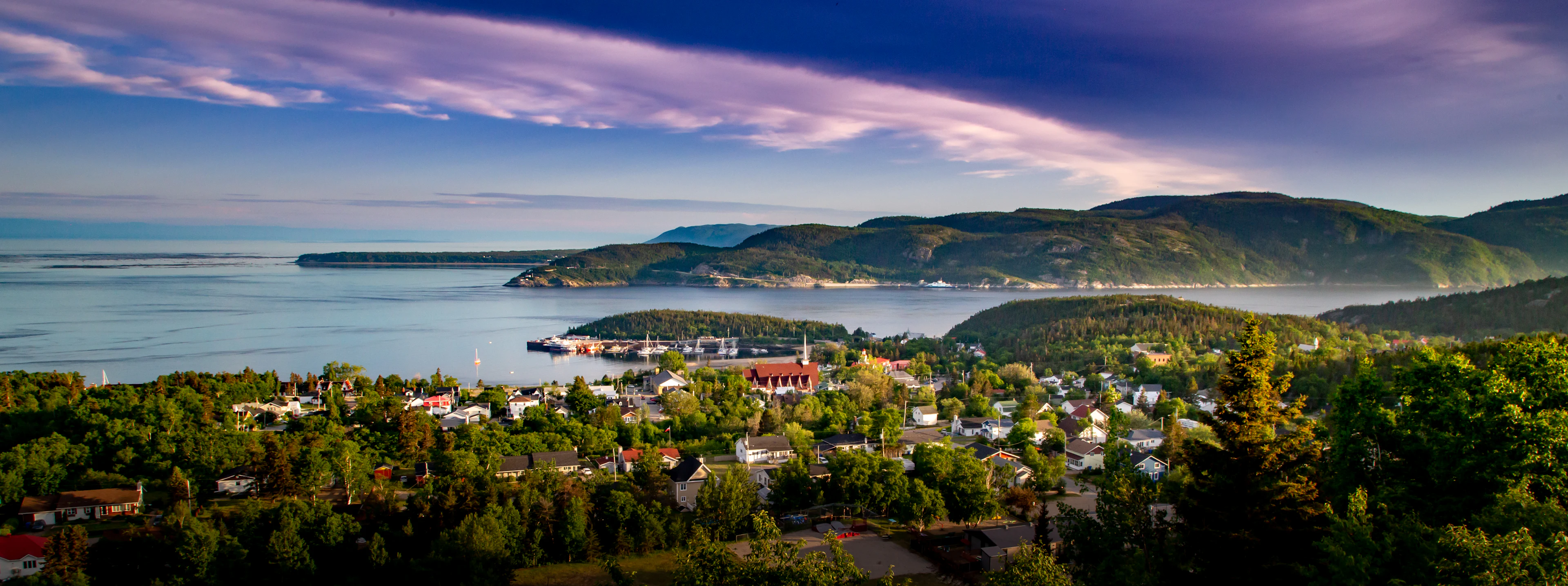 Tadusako miestelis tarp miškingų kalvų prie Saguenay žiočių ir Šv. Lauryno upės, vakaro danguje – rausvi debesys