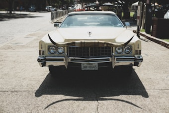 A vintage car with a large set of longhorns mounted on the front. The vehicle is parked on a paved area with buildings and trees in the background. Texas license plate is visible.