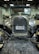 An antique car is positioned inside a dimly lit garage. The vehicle features a shiny chrome grille with round headlights and a license plate that reads '65203 VA, ANTIQUE VEHICLE'. Other cars and automotive equipment are visible in the background.