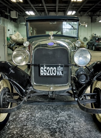 An antique car is positioned inside a dimly lit garage. The vehicle features a shiny chrome grille with round headlights and a license plate that reads '65203 VA, ANTIQUE VEHICLE'. Other cars and automotive equipment are visible in the background.