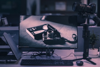 A modern office setup features a widescreen monitor displaying a black and white image of a vintage camera and film. Next to the monitor are a gimbal with a camera attached, a keyboard, a mouse, and a plant in a small pot. The desk is cluttered with cables, and part of a laptop is visible.