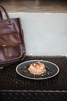 A stack of pancakes topped with almonds and berries sits on a dark plate. The background features a plain, light concrete wall. To the left, a large brown leather bag rests against the wall. The surface beneath is a textured black metal.