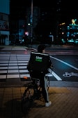 A person riding a bicycle with an Uber Eats delivery bag waits at a crosswalk on a city street at night. The scene is dimly lit with streetlights and neon signs in the background, creating a moody urban atmosphere.