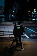 A person riding a bicycle with an Uber Eats delivery bag waits at a crosswalk on a city street at night. The scene is dimly lit with streetlights and neon signs in the background, creating a moody urban atmosphere.