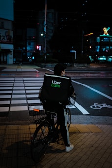 A person riding a bicycle with an Uber Eats delivery bag waits at a crosswalk on a city street at night. The scene is dimly lit with streetlights and neon signs in the background, creating a moody urban atmosphere.