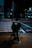 A person riding a bicycle with an Uber Eats delivery bag waits at a crosswalk on a city street at night. The scene is dimly lit with streetlights and neon signs in the background, creating a moody urban atmosphere.
