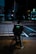 A person riding a bicycle with an Uber Eats delivery bag waits at a crosswalk on a city street at night. The scene is dimly lit with streetlights and neon signs in the background, creating a moody urban atmosphere.