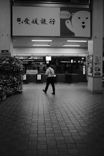 A person walks across a tiled floor in a spacious area, likely part of a train station or public building. Above, a large sign displays Japanese characters alongside a cartoon bear waving. To the side, there is a flower arrangement and some posters on the wall.