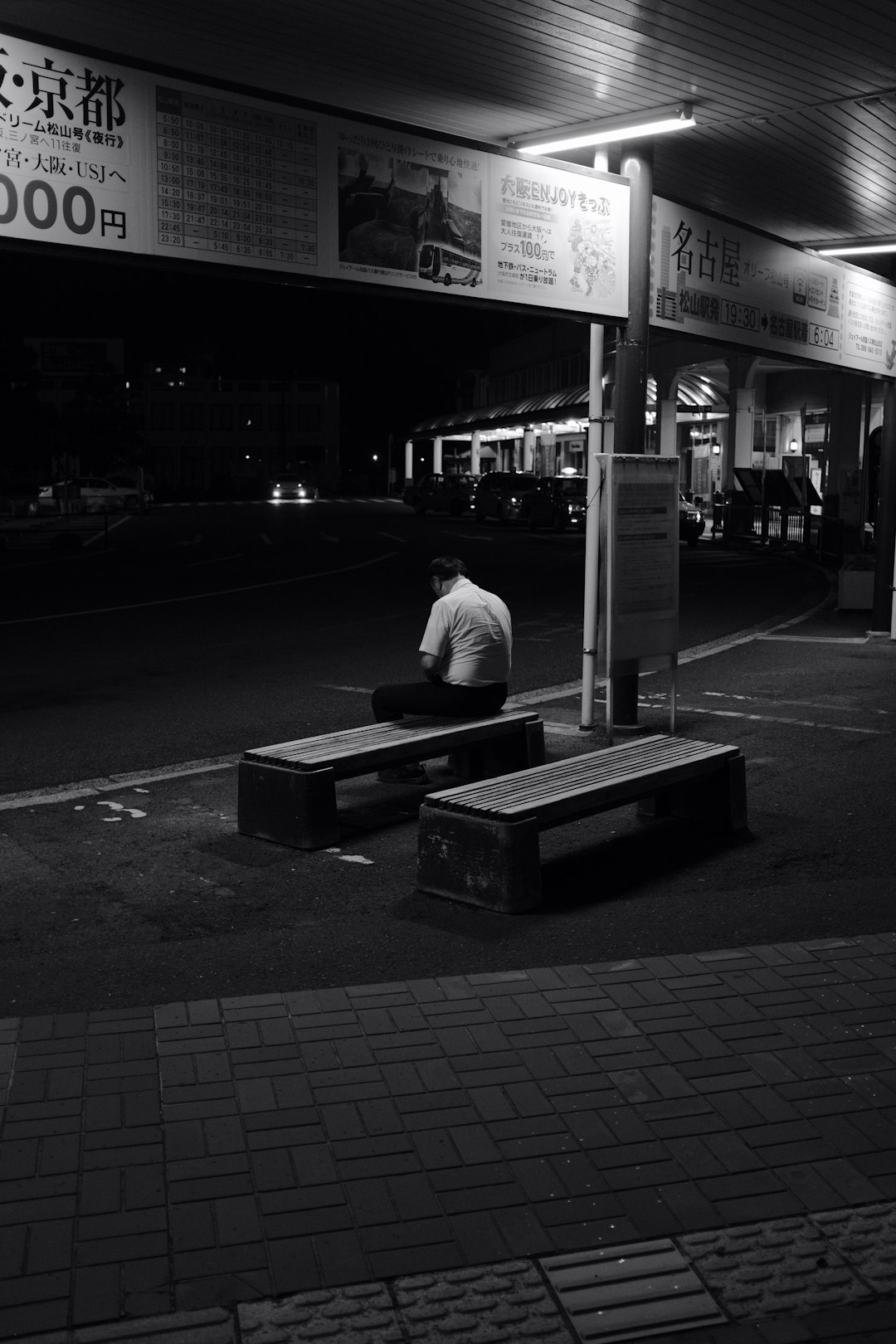 grayscale photo of man in white shirt sitting on bench near store