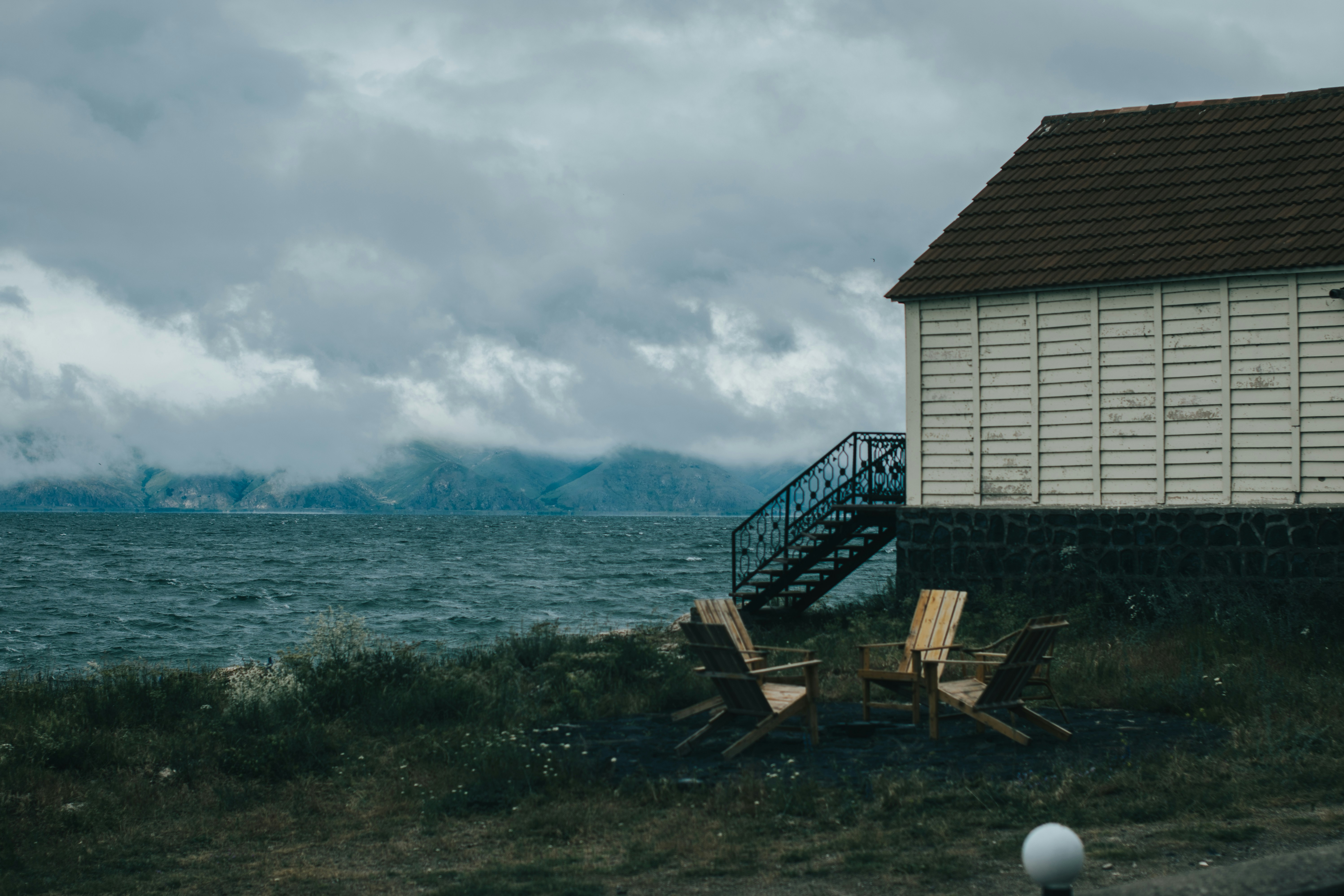 Brown wooden bench on seashore near white and blue house under white clouds during daytime