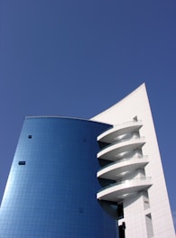 A modern architectural structure featuring a tall building with a curved blue glass facade and distinctive white balconies projecting outward. The sky is clear and bright, providing a crisp backdrop that enhances the building's sleek and clean lines.