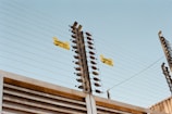 A tall, metallic electric fence with horizontal wires and insulators is seen against a clear blue sky. Yellow warning signs are attached to the fence, indicating it is electrified.