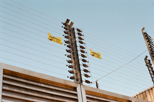 A tall, metallic electric fence with horizontal wires and insulators is seen against a clear blue sky. Yellow warning signs are attached to the fence, indicating it is electrified.