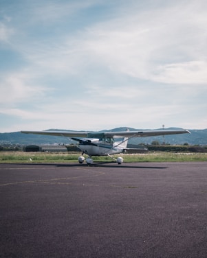 A small propeller airplane is parked on an airstrip with mountains in the background. The sky is partly cloudy, and the surrounding area is grassy with some buildings in the distance.