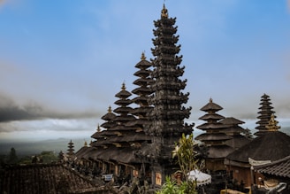An intricate Balinese temple complex with multiple pagoda-style towers, each featuring tiered roofs and ornate carvings. The landscape includes lush greenery and a foggy sky, adding to the serene and mystical atmosphere.