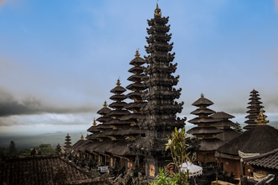An intricate Balinese temple complex with multiple pagoda-style towers, each featuring tiered roofs and ornate carvings. The landscape includes lush greenery and a foggy sky, adding to the serene and mystical atmosphere.
