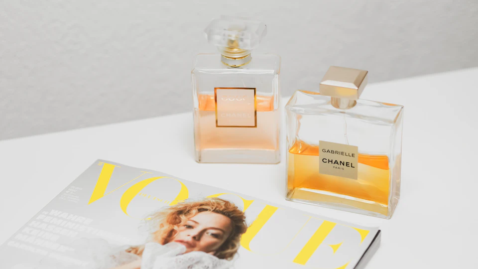 The image presents a scene featuring two perfume bottles, one clear and one gold-colored, placed on top of an open magazine with a picture of a woman's face on it. The clear bottle is positioned slightly to the left of the center, while the gold-colored bottle is located more towards the right side of the image.

The magazine cover displays the name "Vogue" in yellow text at the bottom and features a picture of a woman with blonde hair wearing a white dress.