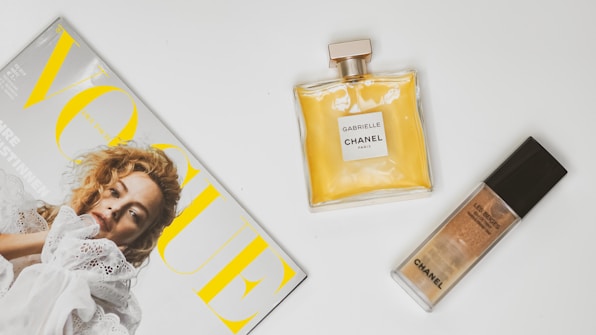 A fashion magazine lies on a white surface, next to a bottle of Chanel perfume and a Chanel foundation. The magazine features a cover with a model wearing a white lace outfit and has prominent yellow lettering.