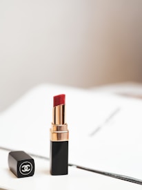 A luxurious red lipstick in a sleek black and gold tube is positioned upright, with its matching black cap lying beside it. The background is softly blurred, creating a minimalistic and elegant aesthetic.