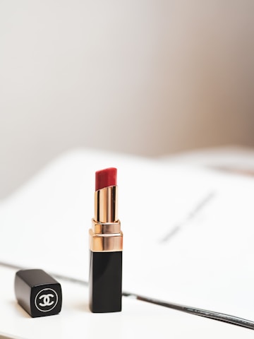 A luxurious red lipstick in a sleek black and gold tube is positioned upright, with its matching black cap lying beside it. The background is softly blurred, creating a minimalistic and elegant aesthetic.