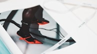 A fashion magazine features an image of vibrant red pointed-toe shoes worn with dark socks. The shoes have a unique design with an artistic heel, and the straps are tied into bows. The page rests open on a surface, alongside a white ribbon with branding printed on it.