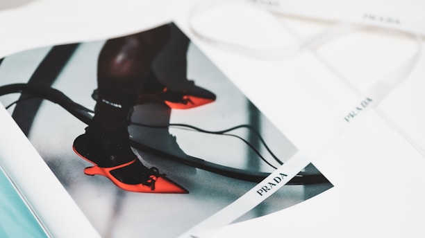 A fashion magazine features an image of vibrant red pointed-toe shoes worn with dark socks. The shoes have a unique design with an artistic heel, and the straps are tied into bows. The page rests open on a surface, alongside a white ribbon with branding printed on it.