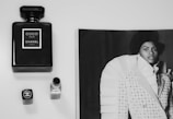 A black and white image featuring a bottle of Chanel Coco Noir perfume, a closed lipstick with a Chanel logo, and a fashion magazine page displaying a model in an elaborate outfit. The objects are arranged on a clean, light surface.