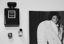 A black and white image featuring a bottle of Chanel Coco Noir perfume, a closed lipstick with a Chanel logo, and a fashion magazine page displaying a model in an elaborate outfit. The objects are arranged on a clean, light surface.