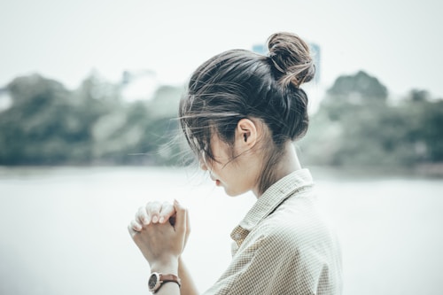 A young person with their hair tied in a bun, wearing a wristwatch and a checkered shirt, appears to be in deep thought or prayer with hands clasped together. The background consists of a blurred outdoor scene with trees, possibly near a body of water.