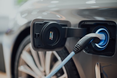 An electric vehicle is being charged, with the plug inserted into the side of the car. The focus is on the charging port and cable, with a blurred view of the car's wheel in the background.