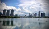 A scenic cityscape featuring a prominent skyline with modern architectural landmarks, such as a unique three-tower structure connected by a rooftop. A large Ferris wheel stands out amidst other skyscrapers. The scene is set against a backdrop of fluffy white clouds in a bright blue sky, reflecting on the calm water surface below.