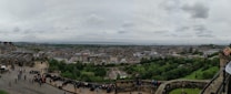 A panoramic view of a city with historical buildings, lush green parks, and busy streets filled with people. The cloudy sky adds a dramatic effect to the cityscape. Visitors are gathered near an ancient stone structure, possibly a castle, observing the view.