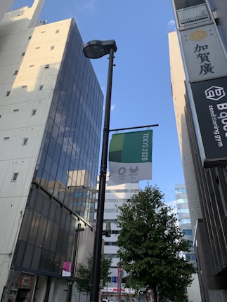 A cityscape view featuring modern high-rise buildings and a street lamp. A banner hanging from the lamp displays logos related to the Tokyo 2020 Olympic and Paralympic Games. A tree is visible in the foreground, and the sky is clear with minimal clouds.