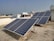 Several solar panels are installed on the rooftop of a building. The panels are angled to face the sun and are supported by a metal framework. The building has a flat roof with a light-colored surface, and other buildings are visible in the background under a clear sky.