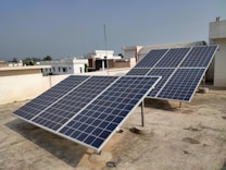 Several solar panels are installed on the rooftop of a building. The panels are angled to face the sun and are supported by a metal framework. The building has a flat roof with a light-colored surface, and other buildings are visible in the background under a clear sky.