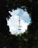 A tall broadcast tower rises prominently into the sky, surrounded by a framing of green foliage. The tower has a modern design with disc-like platforms and an antenna on top. Fluffy white clouds are scattered across the blue sky in the background.