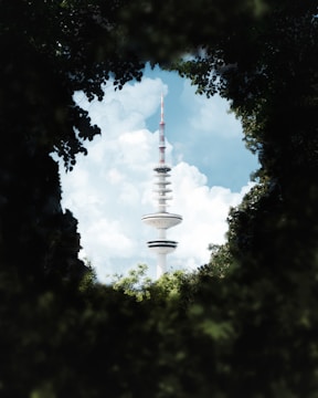 A tall broadcast tower rises prominently into the sky, surrounded by a framing of green foliage. The tower has a modern design with disc-like platforms and an antenna on top. Fluffy white clouds are scattered across the blue sky in the background.