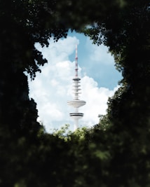 A tall broadcast tower rises prominently into the sky, surrounded by a framing of green foliage. The tower has a modern design with disc-like platforms and an antenna on top. Fluffy white clouds are scattered across the blue sky in the background.